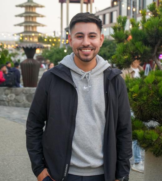Alex is smiling standing outside in front of buildings and trees
