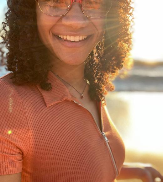 Photo of a person smiling with glasses on with the sun shining behind curly hair
