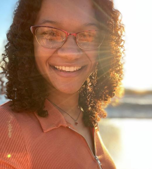 Photo of a person smiling with glasses on with the sun shining behind curly hair