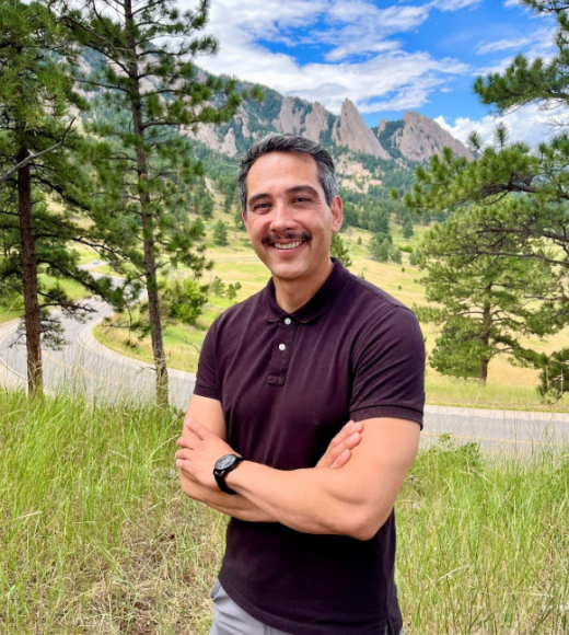Mike is standing outside in front of a scenic mountain view smiling in a polo shirt with crossed arms