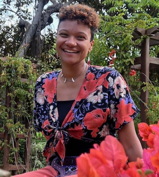 photo of a person with short curly hair smiling outside near flowers and trees