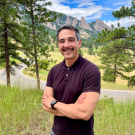 Mike is standing outside in front of a scenic mountain view smiling in a polo shirt with crossed arms 