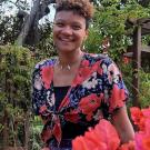 photo of a person with short curly hair smiling outside near flowers and trees